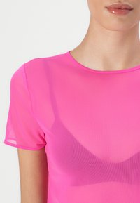 Top leggero e trasparente di colore rosa, con scollatura rotonda e maniche corte, che mostra un reggiseno a contrasto sotto. Finitura liscia e leggermente lucida.