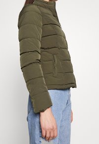 Olivengrøn puffjakke med kort, tætsiddende design, vandrette quiltede sektioner og høj krave. Har en sidelomme med trykknaplukning.