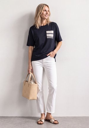 Femme en chemise bleu marine avec poche rayée, pantalon blanc, sandales beiges, tenant un sac à main tressé beige, debout devant un mur clair uni.