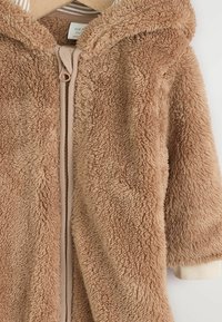 Beige Fleece-Trainingsjacke mit Kapuze. Weiche Textur, gerippte Bündchen und beiger Reißverschluss. Hängt an einem Holzkleiderbügel vor einem weißen Hintergrund.