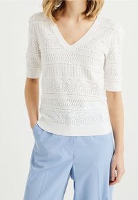 Suéter blanco de punto con escote en V y mangas cortas, que presenta intrincados patrones abiertos y rayas texturizadas. Combinado con pantalones en azul claro.