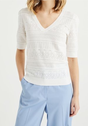 Maglione bianco a maglia con scollo a V e maniche corte, caratterizzato da intricati motivi aperti e strisce testurizzate. Indossato con pantaloni blu chiaro.