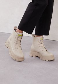 Stivali alla caviglia beige realizzati in morbida pelle scamosciata, con una suola in gomma spessa e un design stringato, dotati di un'etichetta gialla sul tallone e foderati in plaid.