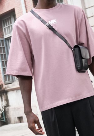 Personne portant un t-shirt rose ample et un pantalon noir, portant un petit sac bandoulière en cuir noir avec une sangle à boucle.