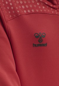 Rød sweatshirt med en tekstureret overdel med et prikket mønster, en glat nederste sektion og et sort logo centreret på brystet.