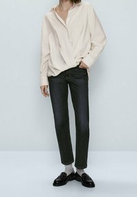 Lichtbeige blouse met knopen, donkere jeans, zwarte instappers, grijze sokken. De blouse is iets oversized met een relaxte pasvorm.