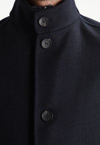 Primer plano de una persona que lleva un abrigo azul marino oscuro con tres botones negros y un cuello alzado sobre un fondo blanco.