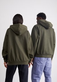 Sweat-shirts vert olive à coupe décontractée, dotés d'une poche kangourou et de poignets côtelés, portés par deux personnes avec des pantalons foncés.
