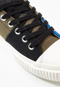 Canvas sneakers med ett mörkgrönt och svart lapptäcke-design, med svarta snörningar, silverfärgade öglor och en vit gummisula.