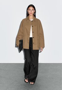 Bruine oversized jas met knopen, gecombineerd met een witte top en zwarte wijde broek. Getextureerde zwarte clutch en open teen schoenen.