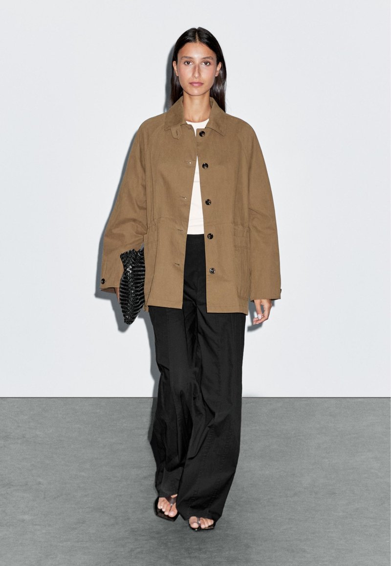 Bruine oversized jas met knopen, gecombineerd met een witte top en zwarte wijde broek. Getextureerde zwarte clutch en open teen schoenen.