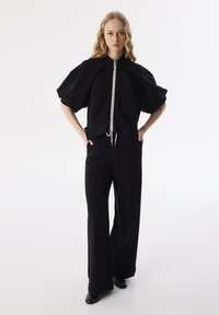 Zwarte oversized top met een hoge kraag en een rits aan de voorkant, gecombineerd met hoge taille zwarte broek, beide in een gladde stofstructuur.