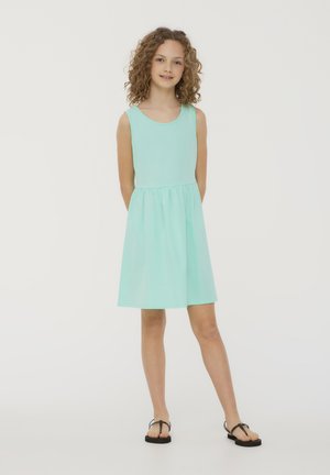 Un vestito senza maniche di colore verde menta con scollatura arrotondata e vita arricciata, realizzato in un tessuto morbido, che arriva sopra il ginocchio. Si indossano sandali neri casual.