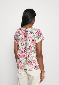 Kwiatowa bluzka z krótkim rękawem na jasnobeżowym tle. Posiada różowo-zielone wzory kwiatowe. Luźny fason, okrągły dekolt, miękka tekstura materiału.