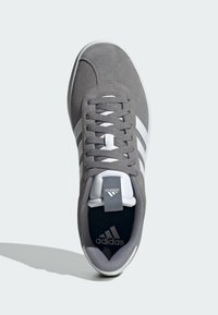Zapatilla de ante gris con tres rayas blancas, cordones texturizados y una lengüeta de marca con interior negro y suela blanca.
