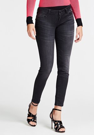 Woman wearing black skinny jeans, a red and black long-sleeve top, and black strappy high-heeled sandals, standing against a white background.