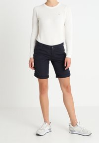 Pantaloni corti blu navy con risvolti, abbinati a una maglietta a maniche lunghe di tessuto leggero. Calzature con accenti bianchi e grigi.