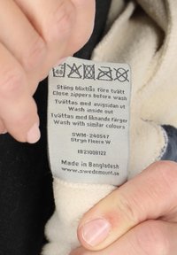 Zorglabel voor een fleece kledingstuk in lichtgrijs. Bevat wasinstructies, verzorgsymbolen en productdetails in het zwart gedrukt.