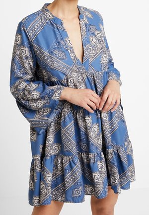 Femme portant une robe bleue à manches longues avec un motif paisley blanc et noir et une taille froncée, les mains jointes près de la taille.