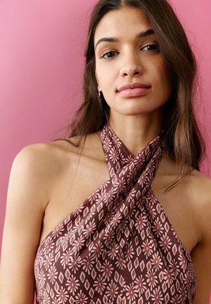 Giovane donna con lunghi capelli castani che indossa un vestito smanicato con collo ad anello e un motivo floreale rosa e marrone, su uno sfondo rosa.