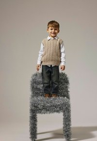 Chaleco de punto gris claro en un niño, camisa blanca abajo, jeans de mezclilla, de pie sobre una silla cubierta de canutillo plateado texturizado.