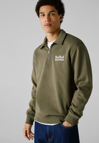 Sudadera de color verde oliva con media cremallera, con dobladillo y puños acanalados, y un logo blanco de "Red Bull RACING" en el pecho.