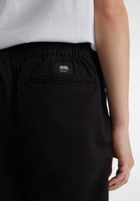 Zwarte Vans-broek met een elastische tailleband, achterzakken en een logopatch. De stof lijkt glad te zijn met een matte afwerking.