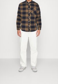 Camisa de franela a cuadros en azul marino, marrón y beige. Presenta cierre de botones y un bolsillo en el pecho. Combinada con pantalones blancos y zapatillas grises.