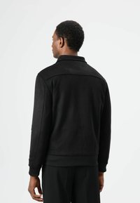 Zwarte suède jas met een kraag, geribbelde manchetten en zoom. Gezien op een model van achteren, met een gladde textuur en minimalistisch ontwerp.