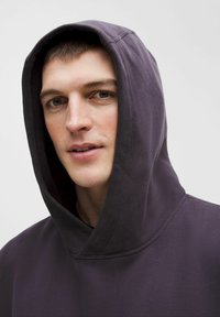 Lilla hættetrøje med en stor, blød hætte og en afslappet pasform. Materialet ligner bomuld og har en glat tekstur samt et minimalistisk design.