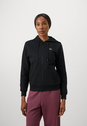 Svart hettejakke med en tettsittende design, snøring i hetten, og grønt logoakcent. Bæres med løse, bordeaux-fargede joggebukser.