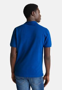 Mladý muž viděný zezadu, oblečený v jasně modrém polo tričku a modrých džínách na jednobarevném bílém pozadí.