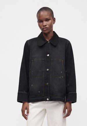 Personne portant une veste noire boutonnée avec de grandes poches avant et des éclaboussures de peinture blanches, associée à un pantalon blanc sur un fond uni.