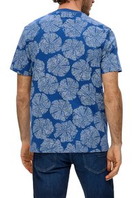 Niebieska koszulka z białym wzorem liści, krótkie rękawy, okrągły dekolt, wykonana z bawełny. Noszona z niebieskimi dżinsami.