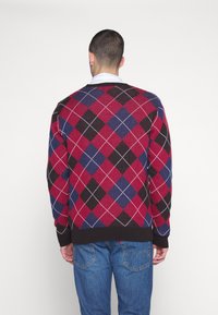 Pull en argyle rouge et marine avec un col et des poignets côtelés, en tissu tricoté texturé ; porté par-dessus un jean bleu.