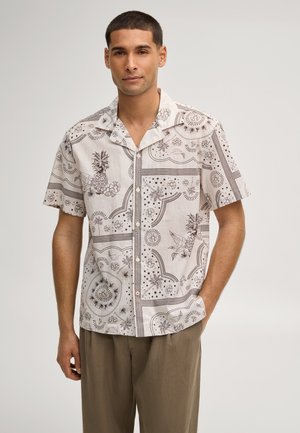 Mężczyzna ubrany w koszulę na guziki z krótkim rękawem, ozdobioną brązowymi botanicznymi i geometrycznymi wzorami, w połączeniu z luźnymi brązowymi spodniami.