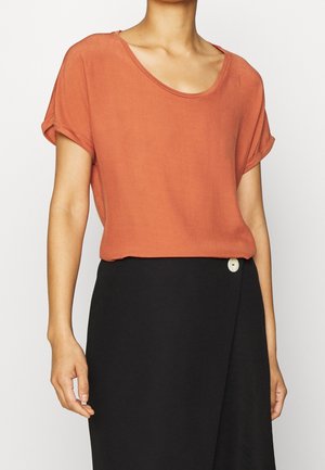 Kobieta w krótkim rękawie, nosząca rdzawy top wsunięty w wysoką taliowaną czarną spódnicę z pojedynczym białym guzikiem.