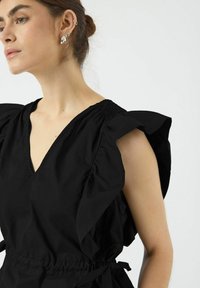 Svart kjole med korte, rysjekantede ermer, V-utringning og rynket detalj ved skuldrene. Snøring i livet, laget av lett stoff.