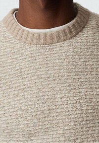Beige strukturierter Strickpullover, der über einem weißen Rundhals-Shirt getragen wird und die obere Brust und den Hals einer Person mit dunkler Haut zeigt.