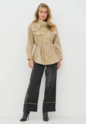 Šviesiai ruda marškinėlių tipo palaidinė su raukinukais, surišta ties liemeniu, derinama su juodomis plačiomis kelnėmis su kontrastingomis siūlėmis ir juodais aukštakulniais bateliais su kniedėmis.