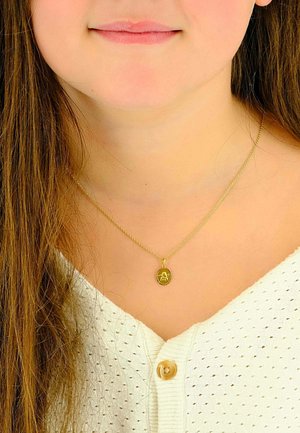 Goldene Anhängerkette mit einem ovalen Charme, der ein detailliertes Design aufweist, an einer dünnen, gedrehten Goldkette. Getragen über einem weißen, strukturierten Oberteil.