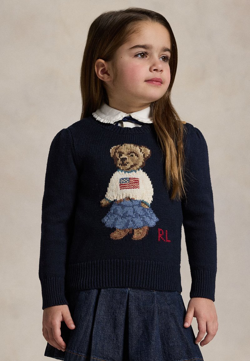 Junges Mädchen schaut zur Seite, trägt einen marineblauen Pullover mit einem Teddybär-Motiv und einen blauen plissierten Rock.