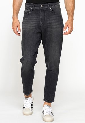 Uomo che indossa jeans neri aderenti e scarpe da ginnastica bianche con strisce nere, in piedi davanti a uno sfondo bianco.