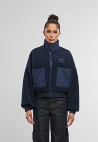 Marineblå kort jacka i fluffig fleece med hög krage, utrustad med tv�å stora framfickor och en texturerad yta.