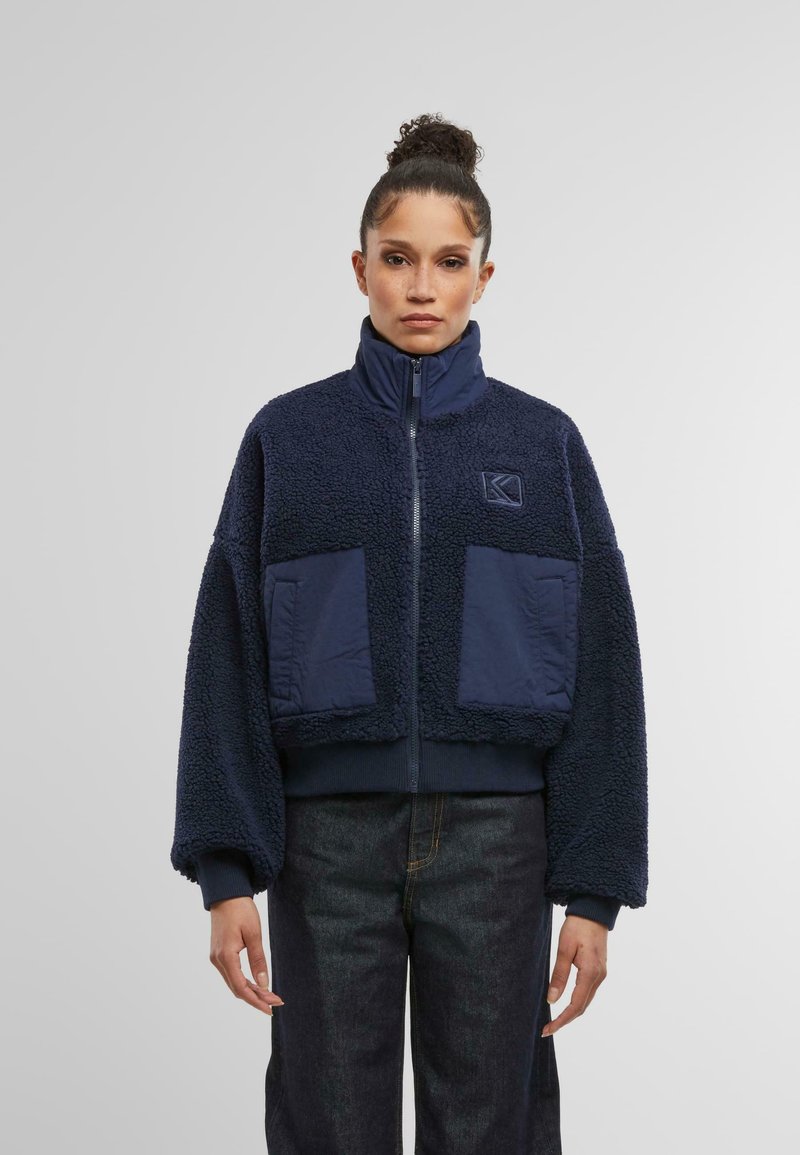 Marineblå kort jacka i fluffig fleece med hög krage, utrustad med två stora framfickor och en texturerad yta.