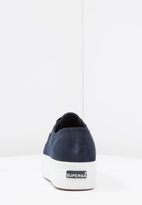 Navy suede sneaker with a white platform sole, featuring a branded tag on the heel and a textured upper with minimal stitching details.