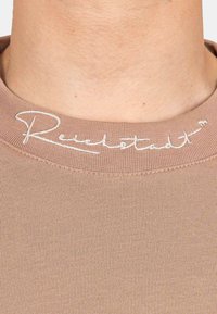 T-shirt marrone aderente con scollatura a coste e logo bianco ricamato sul colletto. Il tessuto sembra liscio e morbido.