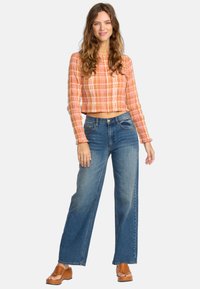 Femme aux cheveux ondulés portant un crop top à carreaux rose-orange, un jean bleu taille haute à jams larges et des sandales à plateforme marron, se tenant sur un fond blanc.