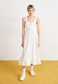 Robe en coton blanche avec un décolleté carré, taille cintrée et ourlet volanté. Associée à des baskets blanches et des chaussettes chevilles sur un sol jaune.