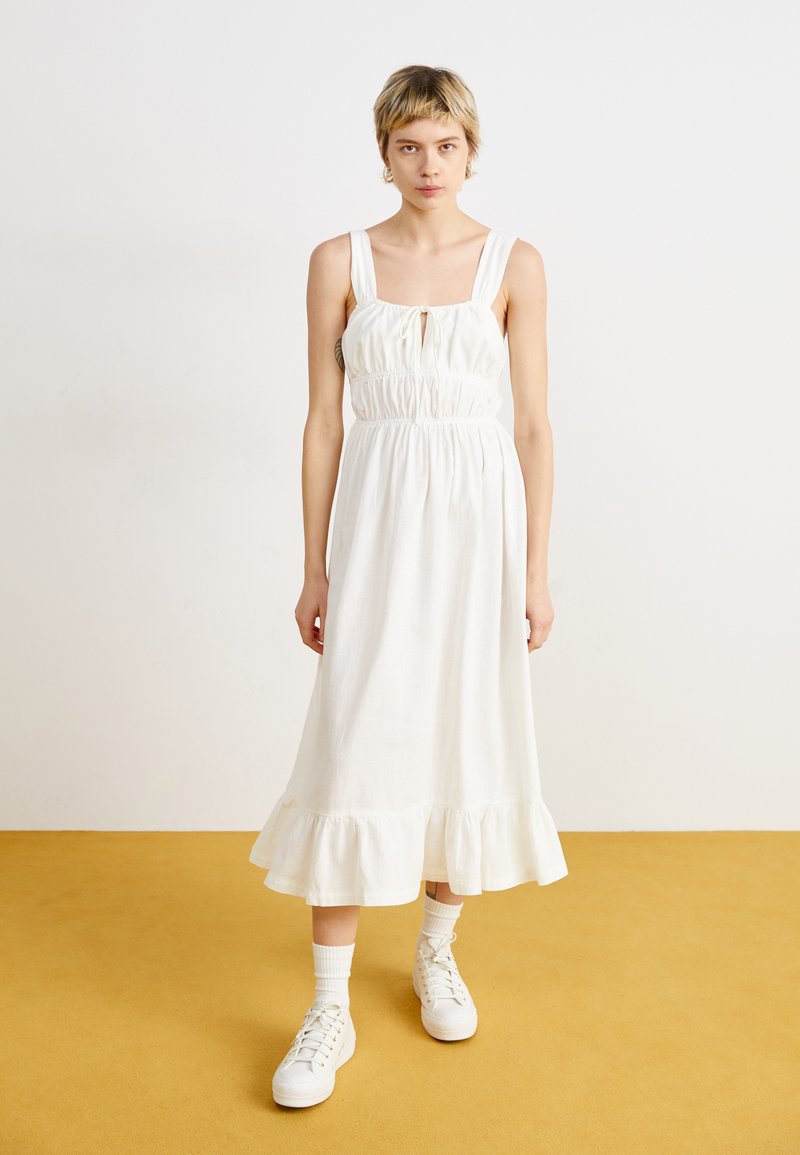 Robe en coton blanche avec un décolleté carré, taille cintrée et ourlet volanté. Associée à des baskets blanches et des chaussettes chevilles sur un sol jaune.